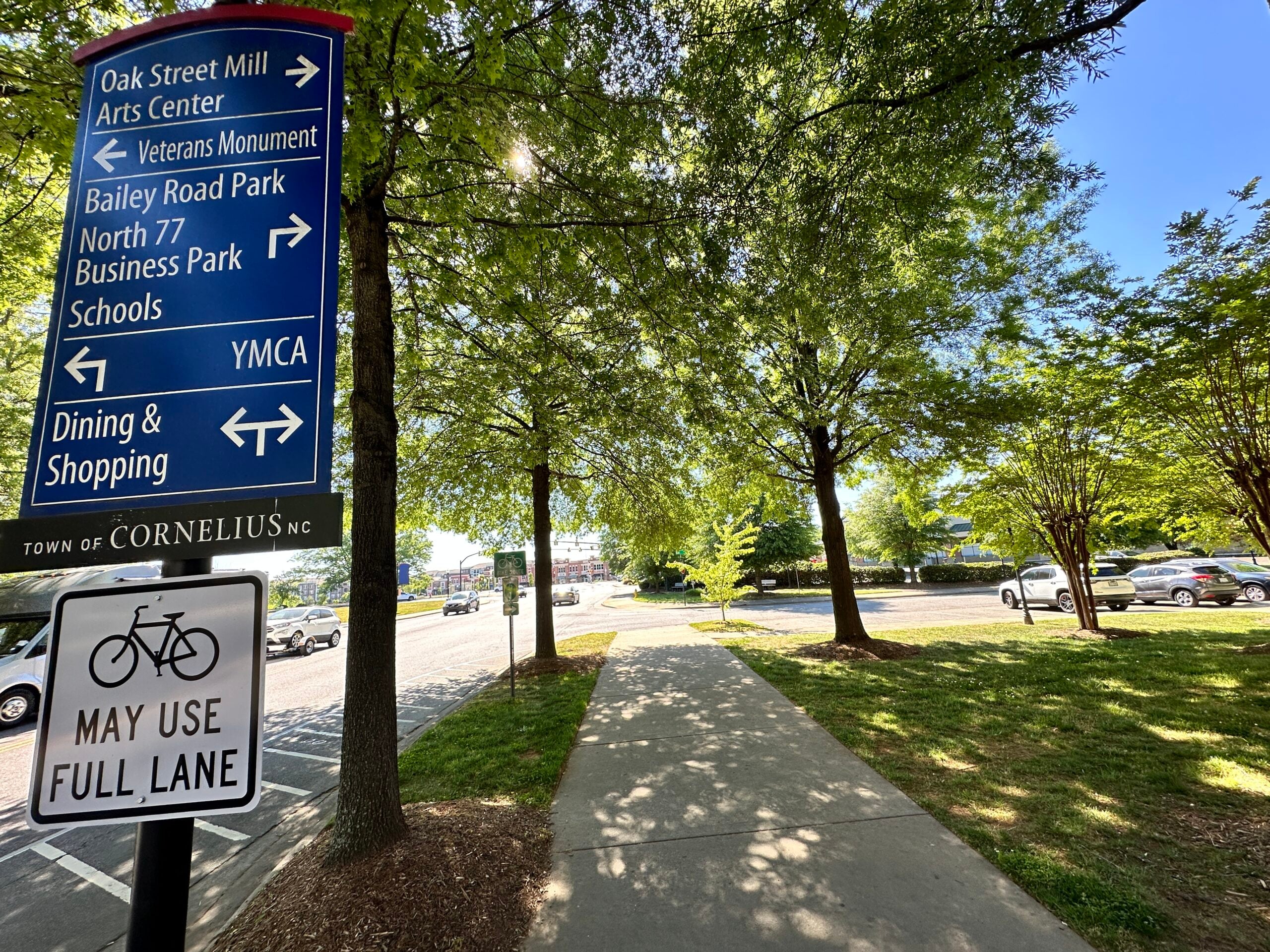 Town of Cornelius wayfinding sign listing Oak Street Mill Arts Center, Bailey Road Park and the YMCA beside a tree-shaded sidewalk